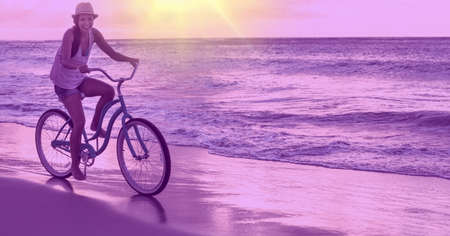 Composition of smiling man riding bicycle on beach by sea with purple tint. holiday, sport, fitness and active lifestyle concept digitally generated image.の写真素材