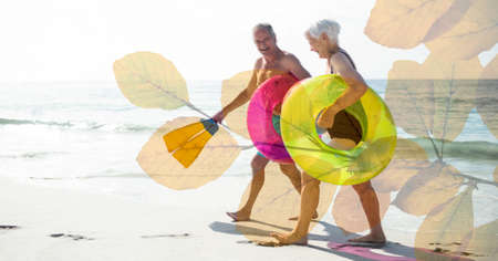 Composition of happy senior couple holding inflatables, smiling on beach and autumn foliage. healthy active retirement lifestyle concept digitally generated image.の写真素材