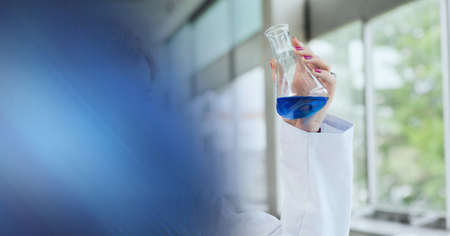 Composition of hand of lab technician holding flask of blue liquid, with blue blurred copy space....