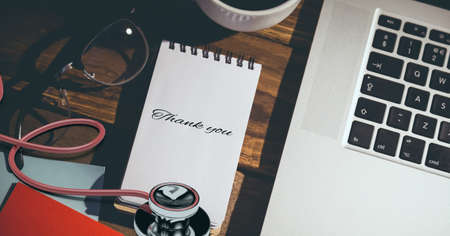 Composition of notebook with thank you note, with laptop, stethoscope and coffee cup on wooden desk. medical services, research and science concept digitally generated image.の写真素材