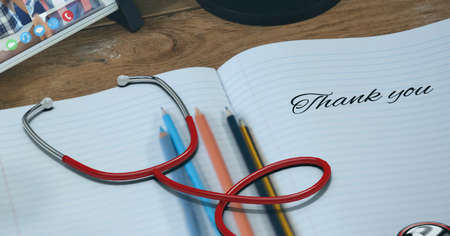 Composition of notebook with thank you note, with stethoscope pencils and smartphone on wooden desk. medical services, research and science concept digitally generated image.の写真素材