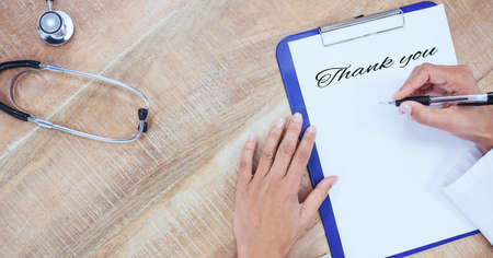 Composition of hands writing thank you note on clipbaord, with stethoscope on wooden desk. medical services, research and science concept digitally generated image.の写真素材