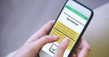 Composition of woman holding smartphone with covid 19 international certificate of vaccination. global coronavirus pandemic, health and covid 19 passport concept digitally generated image.の写真素材