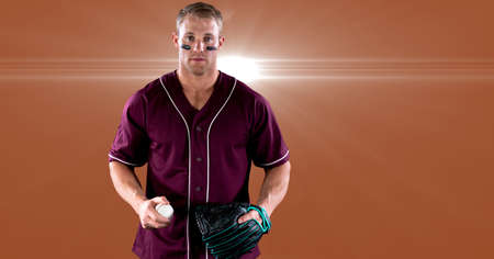 Portrait of caucasian male baseball pitcher holding ball against spot of light on orange background....