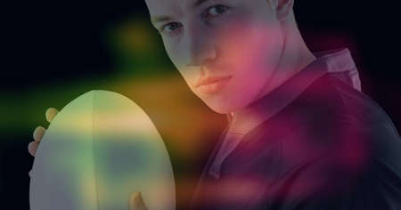Composition of glowing yellow, blue and red blur over male rugby player holding rugby ball, on black. sport and competition digitally generated imageの写真素材