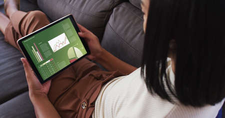 Mixed race woman sitting on sofa using tablet with statistics on screen. communication, data...