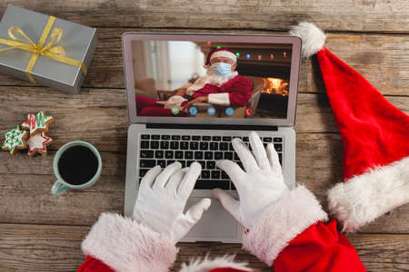 Santa claus on christmas laptop video call with caucasian santa wearing face mask. christmas, festivity and communication technology.の写真素材