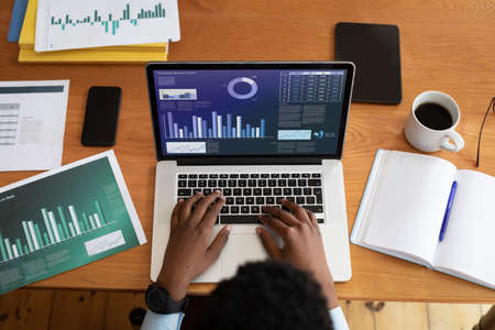 African american businessman sitting at desk using laptop with graph and statistical data on screen....