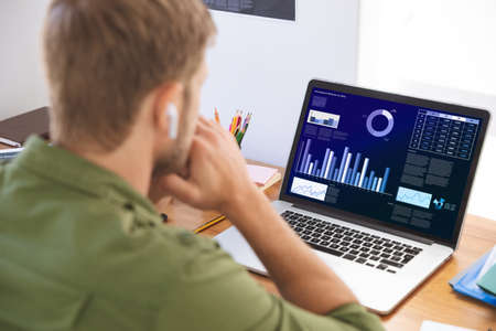 Caucasian businessman sitting at desk using laptop with graph and statistical data on screen. business communication and digital interface technology digital composite image.の写真素材