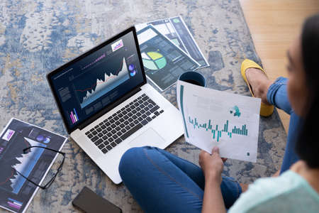 Biracial woman sitting on floor, using computer with statistical data on screen. business communication and digital interface technology digital composite image.の写真素材
