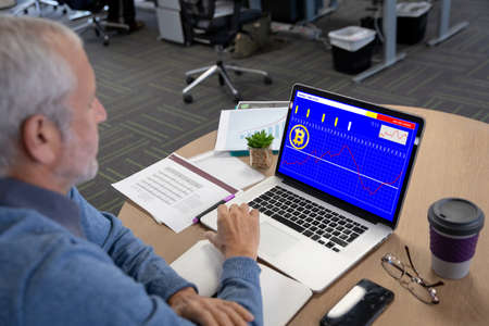 Caucasian senior businessman sitting at desk, using laptop with statistical data on screen. business...