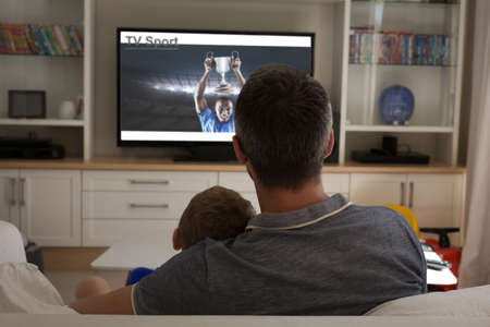 Rear view of father and son sitting at home together watching sport event on tv. sports, competition, entertainment and technology concept digital composite image.の写真素材