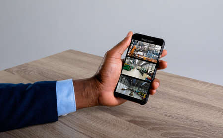 Hand of caucasian man holding smartphone with view of warehouse from security cameras on screen. security, safety, surveillance, and digital interface.の写真素材