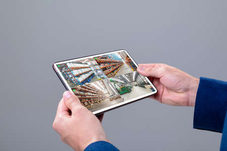 Hands of caucasian man holding tablet with view from security cameras at warehouse on screen....
