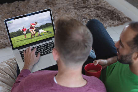 Two diverse male friends at home watching rugby match on laptop. sports, competition, entertainment and technology concept digital composite image.の写真素材