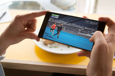 Hands of caucasian man at restaurant watching athletics on smartphone. sports, competition, entertainment and technology concept digital composite image.の写真素材