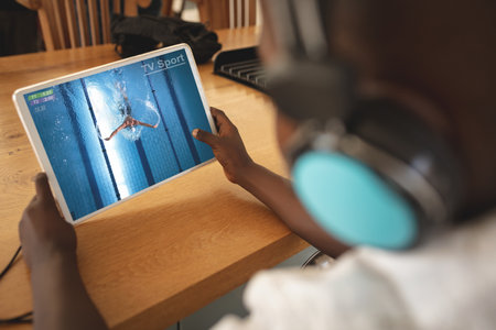 African american boy wearing headphones at home watching swimming competition on tablet. sports,...
