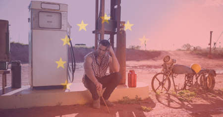 Digital composite image of upset young man sitting at petrol station with european union flag. identity and lifestyle.の写真素材