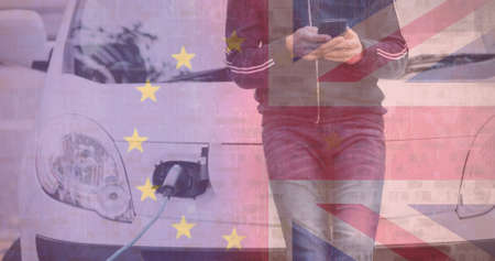 Man using smartphone waiting at electric charging station with european union and british flags. digital composite of environmental conservation, transportation and identity.の写真素材