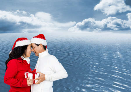 Composite image of asian senior couple in santa hats holding gift against blue sky over the sea....