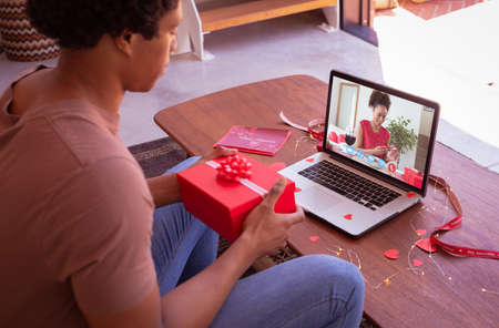 African american young man showing valentine day gift to girlfriend on video call through laptop. unaltered, online dating, video call and distant valentine day celebration.の写真素材