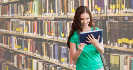 Smiling caucasian teenage girl reading book while standing against bookshelf in library. digital...