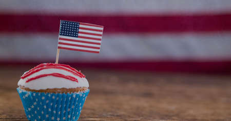 Miniature american flag over a cup cake on wooden surface against american flag in background. american independence and patriotism conceptの写真素材