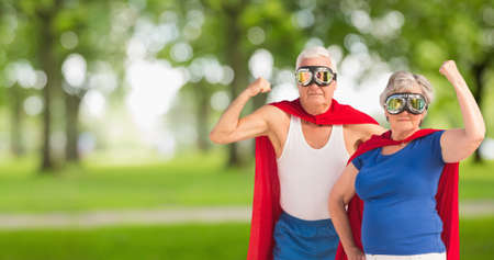Portrait of caucasian senior couple wearing red capes and masks flexing muscle against trees in park. love, togetherness, superhero, power, fantasy, unaltered, senior citizens, retirement, awareness.の写真素材