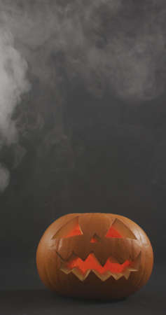 Vertical image of lit carved halloween pumpkins with clouds of smoke on grey background. halloween, tradition and celebration concept.の写真素材