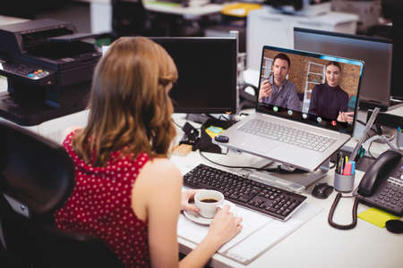 Caucasian businesswoman with coffee cup on desk video conferencing with coworkers over laptop. drink, unaltered, technology, teamwork, meeting, discussion, business, occupation and office concept.の写真素材
