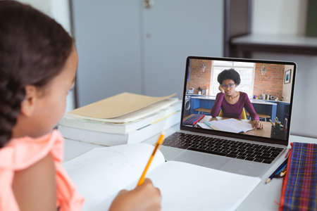 Multiracial girl writing notes while female teacher teaching over video call on laptop at home....