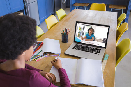 African american girl writing in notebook while taking notes from friend though video call on...