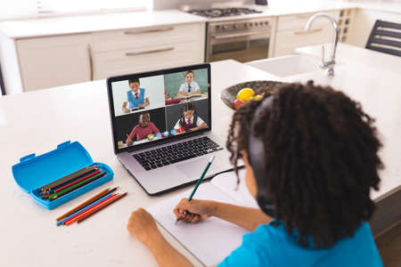 African american boy with curly hair writing notes while learning from online class over laptop....