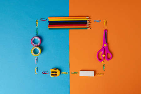 Composition of frame made of crayons, scissors and other items on blue and orange surface. School...