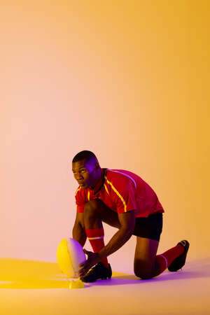 African american male rugby player crouching with rugby ball over pink lighting. Sport, movement, training and active lifestyle concept.の写真素材