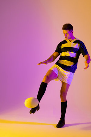Caucasian male rugby player kicking rugby ball over pink lighting. Sport, movement, training and active lifestyle concept.の写真素材