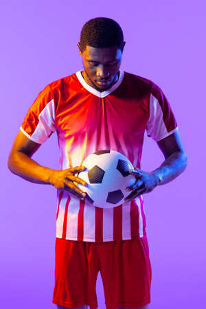 African american male soccer player with football over neon pink lighting. Sport, movement, training and active lifestyle concept.の写真素材