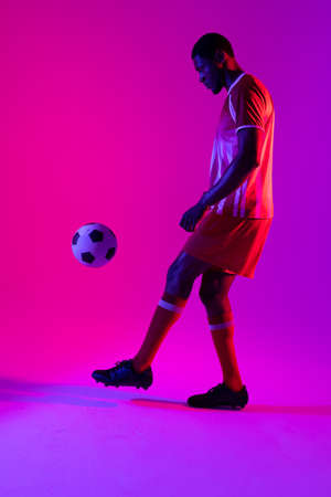 African american male soccer player with football over neon pink lighting. Sport, movement, training and active lifestyle concept.の写真素材