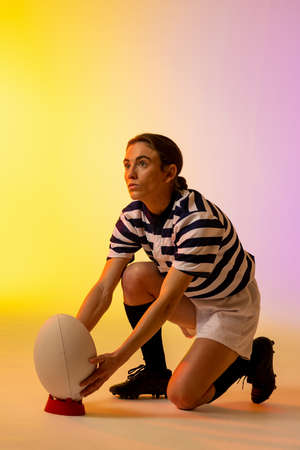 Caucasian female rugby player crouching with rugby ball over neon pink lighting. Sport, movement, training and active lifestyle concept.の写真素材