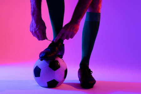 Caucasian male soccer player with football tying shoes over neon pink lighting. Sport, movement, training and active lifestyle concept.の写真素材