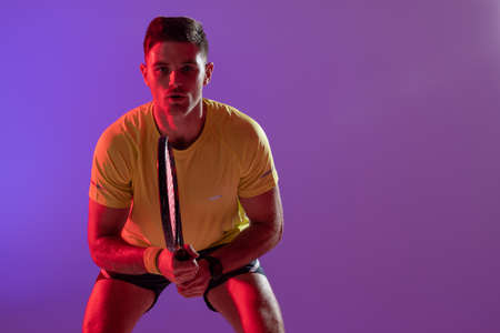 Portrait of caucasian male tennis player with tennis racket over neon pink lighting. Sport,...
