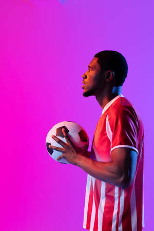 African american male soccer player with football over neon pink lighting. Sport, movement, training and active lifestyle concept.の写真素材