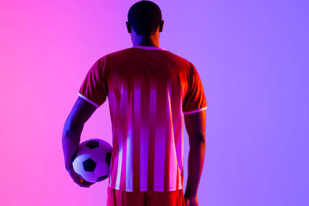 African american male soccer player with football over neon pink lighting. Sport, movement, training and active lifestyle concept.の写真素材