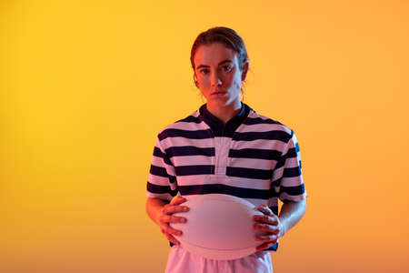 Portrait of caucasian female rugby player with rugby ball over neon yellow lighting. Sport, movement, training and active lifestyle concept.の写真素材