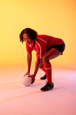 Portrait of african american female rugby player with rugby ball over neon yellow lighting. Sport, movement, training and active lifestyle concept.の写真素材