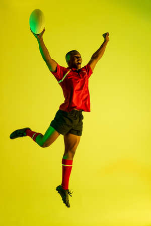 Happy african american male rugby player raising hands over yellow lighting. Sport, movement, training and active lifestyle concept.の写真素材