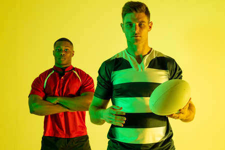 Portrait of diverse male rugby players with rugby ball over yellow lighting. Sport, movement, training and active lifestyle concept.の写真素材