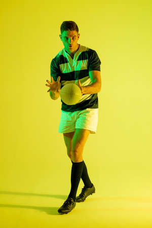 Caucasian male rugby player catching rugby ball over yellow lighting. Sport, movement, training and active lifestyle concept.の写真素材