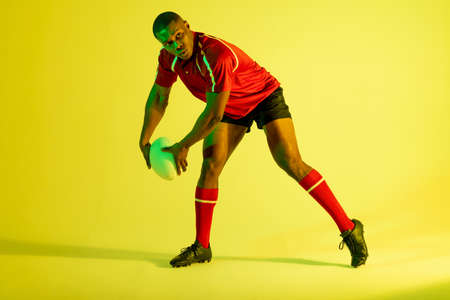 African american male rugby player with rugby ball over yellow lighting. Sport, movement, training and active lifestyle concept.の写真素材