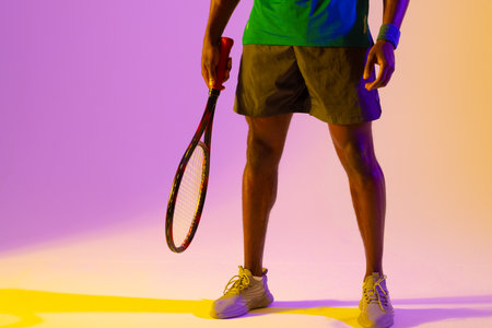 Image of low section of african american male tennis player in violet and yellow neon lighting....